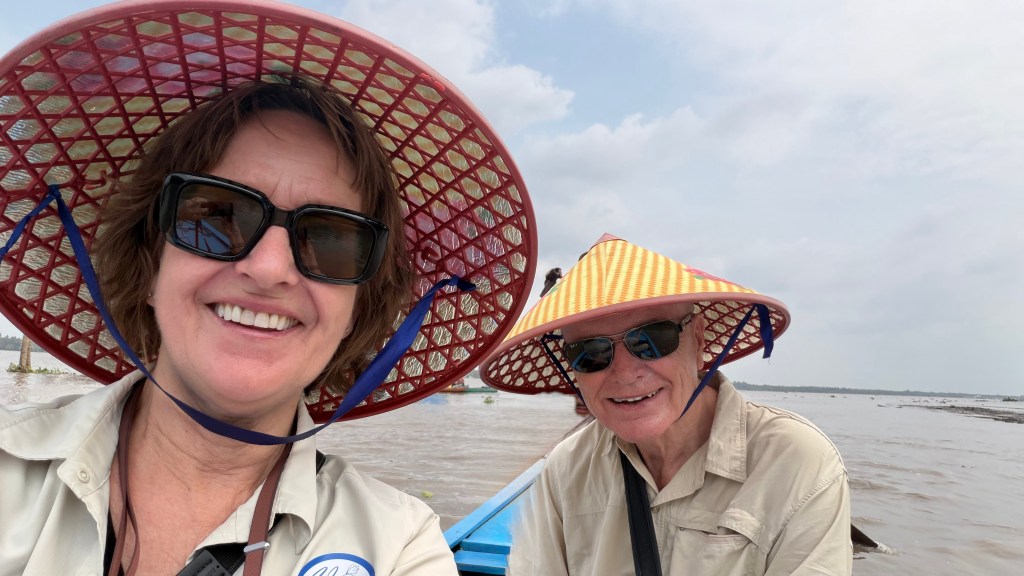 A smiling woman and man wearing traditional conical hats and sunglasses, sitting in a boat on a river. 